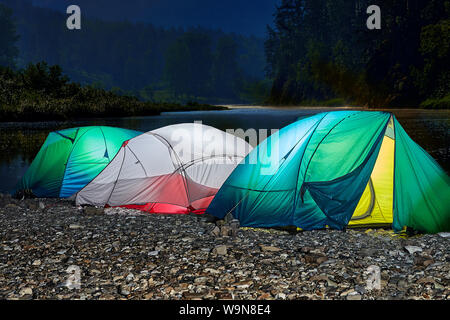 Three multi-colored tourist tents are illuminated from inside, set in nature, near the river, under a starry night sky. Stock Photo