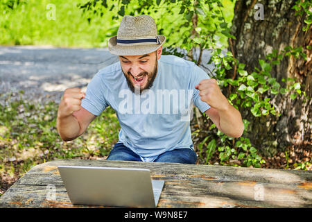 Young bearded man in a hat is sitting near a laptop in nature, emotionally rejoices looking at the screen. Stock Photo