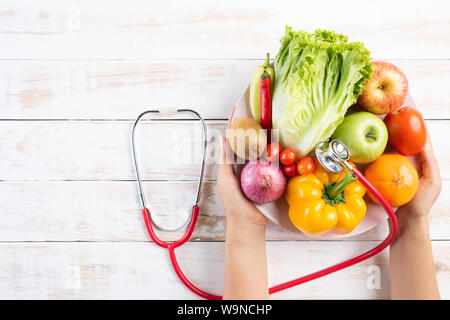 Healthy lifestyle, food and nutrition concept. Close up doctor woman hand holding plate of fresh vegetables and fruits with stethoscope lying on white Stock Photo