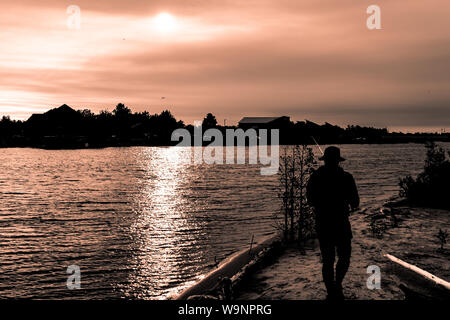 Silhouette of guy in hat fishing in river on pink sunset Stock Photo