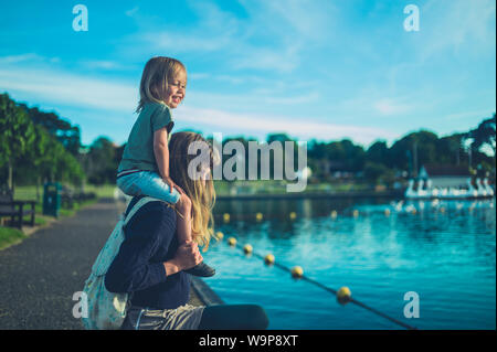 A little toddler is sitting on his mother's shoulders by a lake in a park at sunset Stock Photo