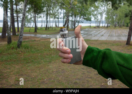 Vaporizer in hand. Man holds vape e-cigarrete device with thumbs-up gesture on nature background. Concept of alternative smoking as positive or negati Stock Photo