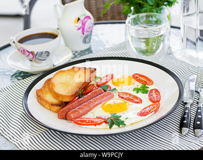 English breakfast on summer terrace: fried eggs, sausage, tomatoes, and toasts. Cup of coffee. Close up of hotel breakfast. Stock Photo
