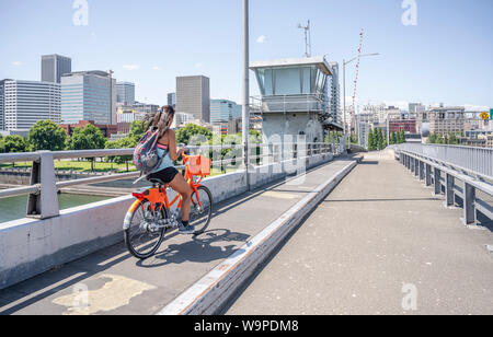A young girl with her hair and backpack rides an rented orange bike over the bridge and simultaneously flows on the phone, representing the lifestyle Stock Photo