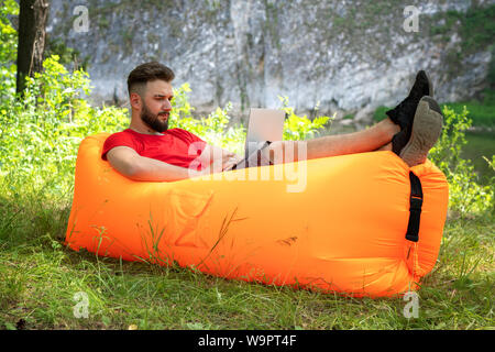Young bearded businessman with laptop on his knees is sitting  on the orange inflatable sofa on the grass in forest. Stock Photo
