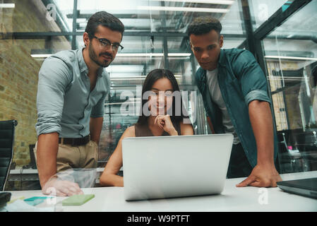 Great results! Group of three young employees looking at screen of laptop and smiling while working in modern office. Job concept. Business people. Workplace Stock Photo