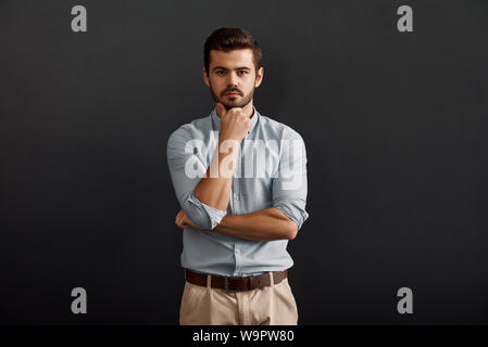 Thinking about business. Serious young bearded man looking at camera and touching his chin while standing against dark background. Work concept. Studio shot Stock Photo