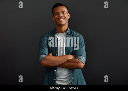 Happy to work here. Cheerful and young afro american man looking at camera with smile and keeping arms crossed while standing against dark background. Business people. Work concept Stock Photo