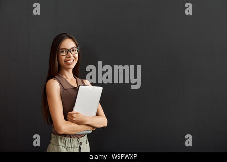 Using modern technologies. Young business woman in casual wear and eyeglasses holding laptop and looking at camera with smile while standing against dark background. Business people Stock Photo