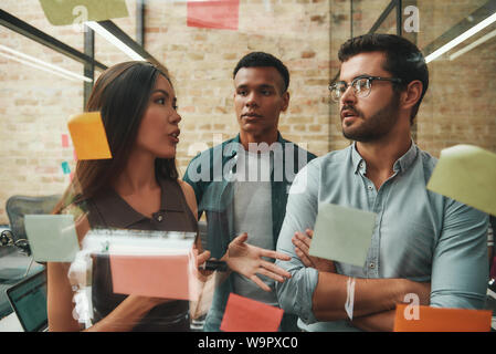 Kanban board. Cheerful male and female colleagues planning working process and using colorful stickers while standing in front of glass wall. Teamwork. Project management Stock Photo