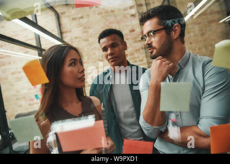IT team. Cheerful male and female colleagues planning working process and using colorful stickers while standing in front of glass wall. Kanban. Project management Stock Photo