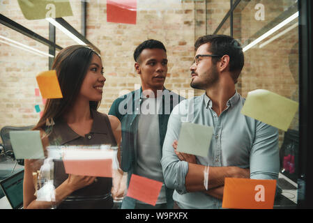 Multicultural team. Cheerful male and female colleagues planning working process and using colorful stickers while standing in modern office. Teamwork. Project management Stock Photo