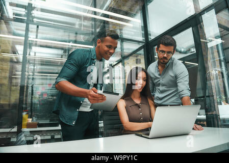 Successful team. Group of three young and cheerful employees using modern technologies and discussing new project while working in modern office. Job concept. Workplace Stock Photo