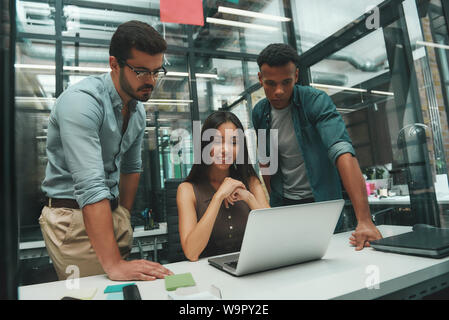 Project team. Group of three young employees looking at screen of laptop and discussing new project while working in modern office. Job concept. Business people. Workplace Stock Photo