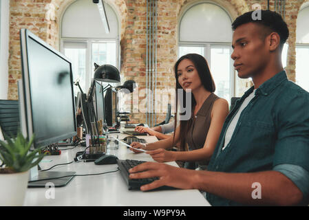 Discussing new business strategy. Two young people in casual clothes looking at computer monitor and talking about something while sitting at desk in modern open space. Job concept. Workplace. Teamwork Stock Photo