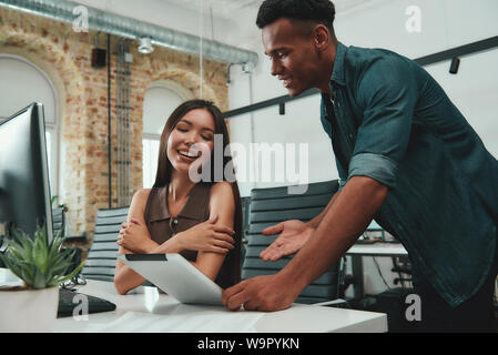 Good news! Young afro american man in casual clothes showing data on his digital tablet and discussing new project with his beautiful female coworker. Business people. Workplace Stock Photo