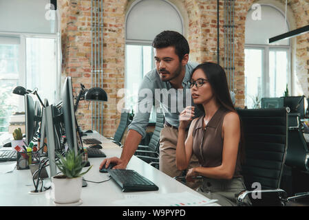 Project team. Two young colleagues in casual wear looking at computer monitor and discussing results while working in modern office. Workplace. Business concept. Stock Photo