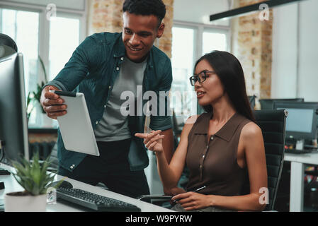 Happy to work together Young afro american man in casual clothes showing something on his digital tablet and discussing new project with his beautiful female coworker. Workplace. Business concept. Stock Photo