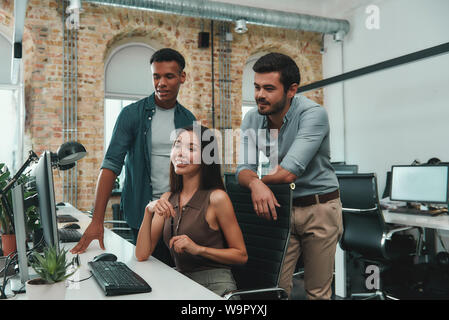 Business professionals. Group of young and cheerful three people discussing new project and looking at computer while working in modern office. Business people. Workplace Stock Photo