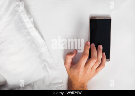 Man trying to snooze the alarm in the morning. Hand touching the smartphone lying in the bed near the pillow. Stock Photo