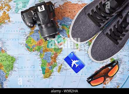 Top view of camera, airplane luggage tag, sunglasses and sneakers on a map of the world. These are my travel essentials for a trip around the world or Stock Photo
