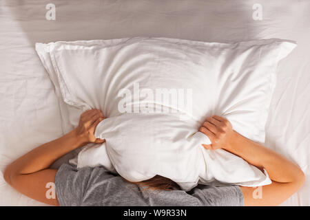 Woman lying in bed and covering her head with pillow. Struggling from insomnia, noise or early morning get up. Stock Photo