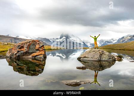 Hiker stands on rocks in the lake and stretches his arms into the air, Lake Stellisee, Matterhorn, cloudy, Valais, Switzerland Stock Photo