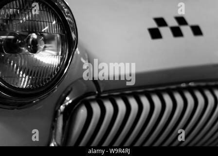 monochrome fragment of the front part of a vintage taxi car, focus on the headlight, checker emblem and radiator in blur Stock Photo