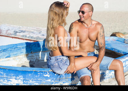 Young adult couple having fun at sandy beach sitting on old rowboat Stock Photo