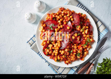 Garbanzos fritos, hot Chickpea stew with sliced chorizo, ham, tomatoes and spices on a white plate on a white concrete table, spanish cuisine, view fr Stock Photo