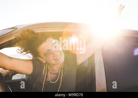 Woman enjoying a beautiful vacation sunny day sitting in a car with raised arms outside the window. Portrait of a smiling girl with trendy sunglasses Stock Photo