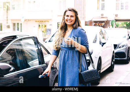 Beautiful blonde pregnant woman opens the driver's door of her car. Stock Photo