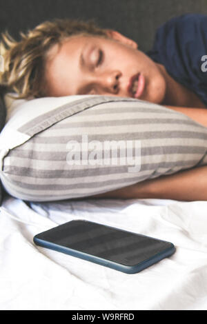 Young boy sleeping with hands under the pillow near the cellphone. Beautiful child sleeps alone in the bed dreaming about online games. Technology add Stock Photo
