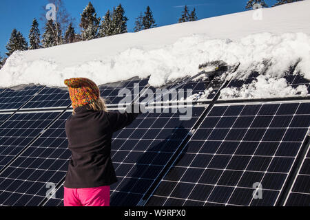 Woman pushing snow off solar panels in winter. If snow covers panels, they can’t produce power. Private small home house on the background. Stock Photo