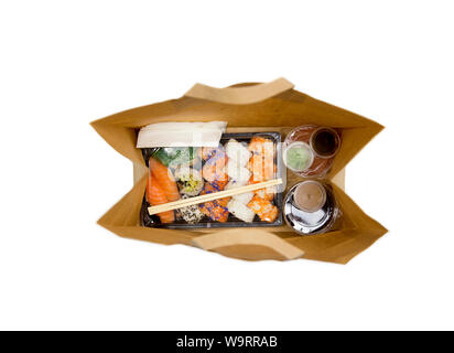 Sushi to go concept. Top view of takeaway box with sushi rolls and various sauce cups in brown paper bag. Isolated on white, studio shot. Stock Photo