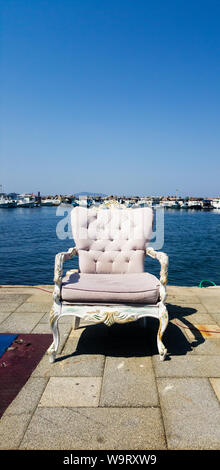 An armchair with baroque decorations standing on cement in the foreground. A fisher port, boats and the Mediterranean sea are seen at the background, Stock Photo