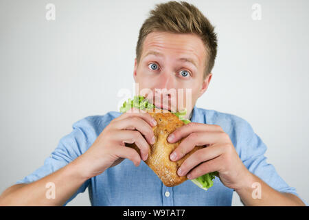 young handsome hungry man biting fresh sandwich with salad leaf holding it with both hands on isolated white background. Stock Photo