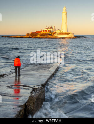 A man stands on the St Mary's Island Causeway while the setting sun illuminates St Mary's Lighthouse on the coast of Whitley Bay in Tyneside. Stock Photo