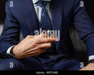 A man in a classic suit sits on a black leather sofa and shows off an expensive petrol lighter. Boss style. Nervous waiting. Criminal authority. Gold Stock Photo