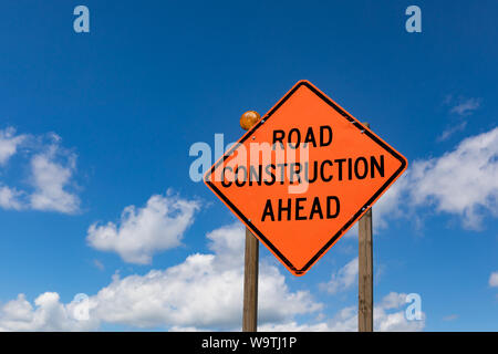 Closeup of orange road construction ahead sign and warning light on sign posts with blue sky and white clouds in background Stock Photo
