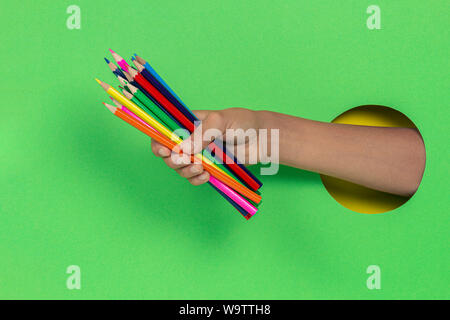 Kid holding colored pencils in hand through hole on light green background Stock Photo