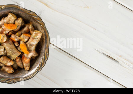 Lot of whole lot of slices of bright turmeric rhizome copyspace on right in old iron bowl flatlay on white wood Stock Photo