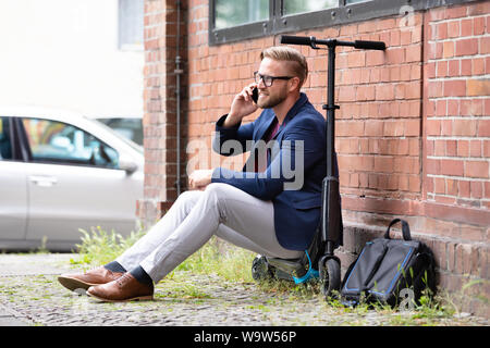 A Handsome Businessman Sitting On Electric Scooter Using Cellphone Near Brick wall Stock Photo