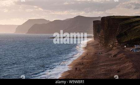 People walking and fishing on Hive Beach at Burton Bradstock are silhouetted against the evening sun on Dorset's Jurassic Coast. Stock Photo