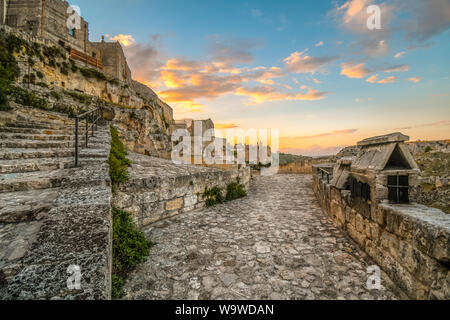 The ancient stone path around the prehistoric city of Matera, Italy, with the sassi and Convent of Saint Agostino in the distance as the sun sets Stock Photo