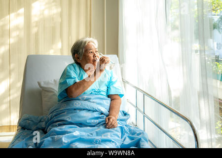 Asian elderly woman sitting and holding glass of water in bed at hospital. Stock Photo