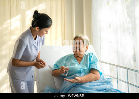 Asian nurse giving medication and glass of water to senior woman at hospital ward. Medicine, age, health care and people concept Stock Photo
