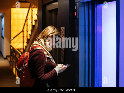 Side view of woman working on mobile phone waiting for the elevator in modern hotel - blue lgiht coing from the oepn door Stock Photo