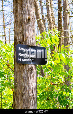 A bilingual trail sign nailed to a tree warns hikers to stay on the designated pathway in both English and French. Stock Photo