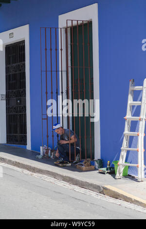 A house painter works on the iron door shutters with all his tools laid by the painted wall in sunny afternoon, Old San Juan, San Juan, Puerto Rico. Stock Photo